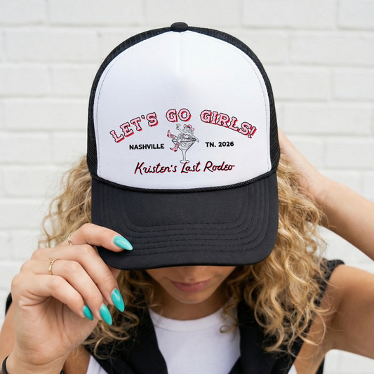 Person wearing a black and white cap with "Let's Go Girls" text and cowgirl bride in a martini glass graphic, against a white brick wall.